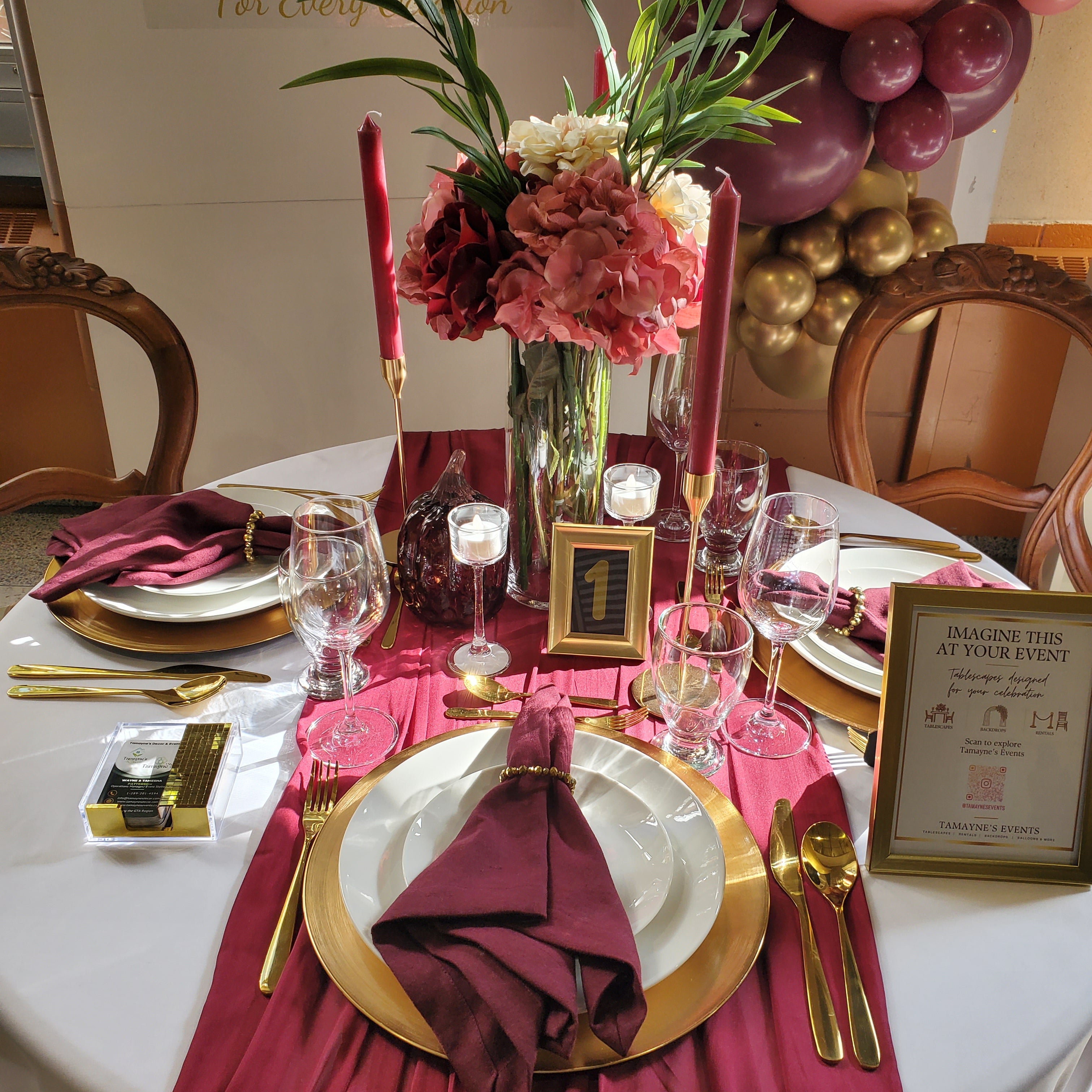A styled round event table set with white tablecloths, burgundy napkins, gold chargers and cutlery, clear wine glasses, a floral centerpiece, red taper candles, and a gold number 1 sign. Chairs and balloons are visible in the background.