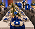 A long banquet table set for a birthday celebration, featuring white tablecloths, blue and white floral centerpieces, blue napkins folded like tuxedo shirts, silverware, and blue draped fabric runners. Background decorations include a 'Happy Birthday 80' sign and tuxedo-themed decor.