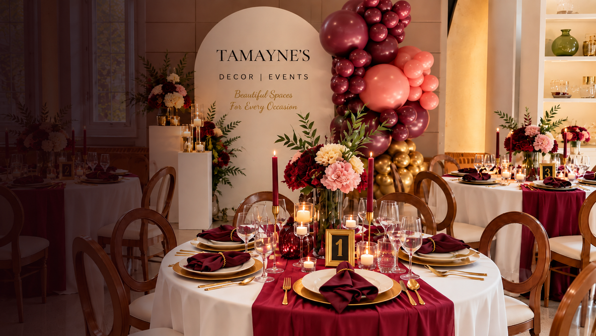 A styled event reception table setup in a banquet hall, featuring round tables with white tablecloths, deep burgundy table runners, gold-rimmed plates, folded burgundy napkins, and floral centerpieces. A large arched backdrop reads 'TAMAYNE'S DECOR EVENTS' with pink, burgundy, and gold balloon garlands rising behind the table.