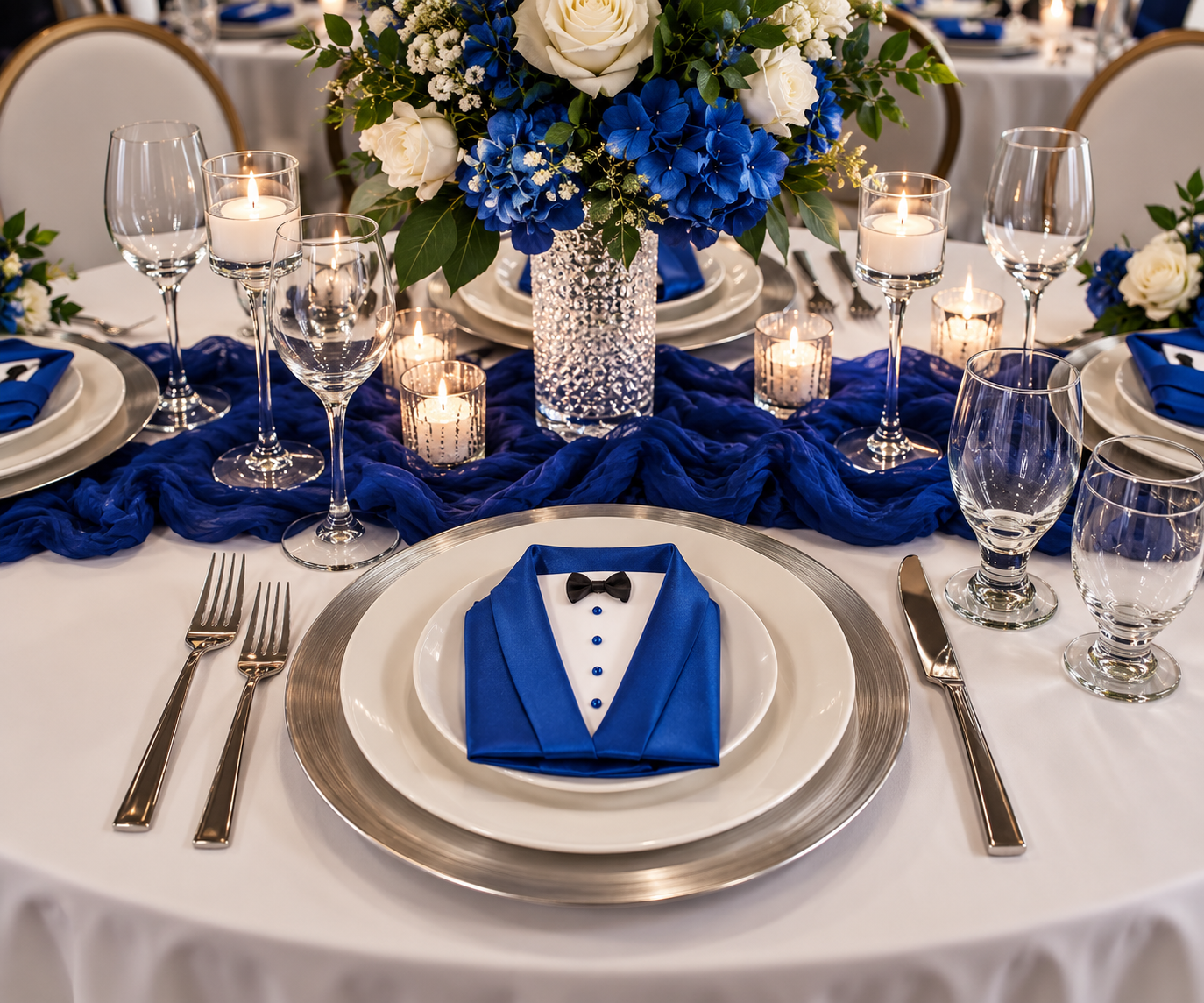 A formal event table setting featuring a blue and white place setting with a folded napkin styled as a tuxedo jacket with a bow tie. The table is decorated with a blue and white floral centerpiece, blue draped fabric, clear glassware, and lit candles.