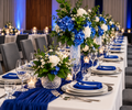 A long banquet table set for guests with white tablecloths, blue napkins, layered white plates, clear wine glasses, and floral centerpieces featuring blue hydrangeas, white roses, and green foliage.