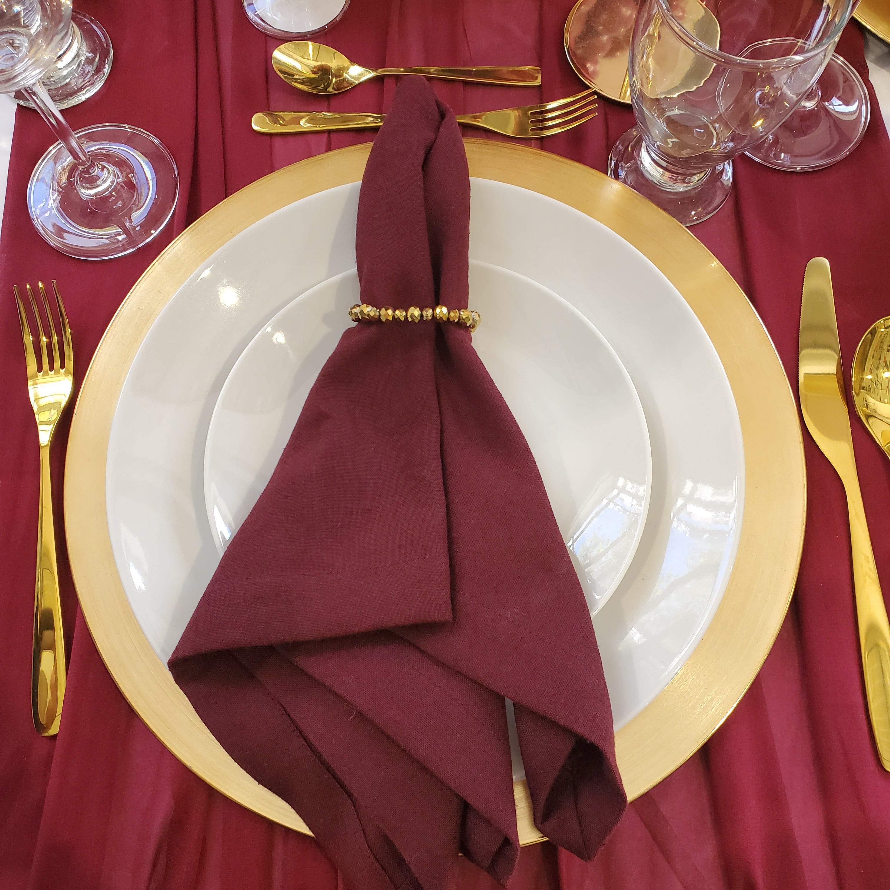 A styled table setting featuring a white dinner plate with a wide gold charger plate, a folded burgundy napkin secured by a gold beaded ring, gold flatware, and red tablecloth.