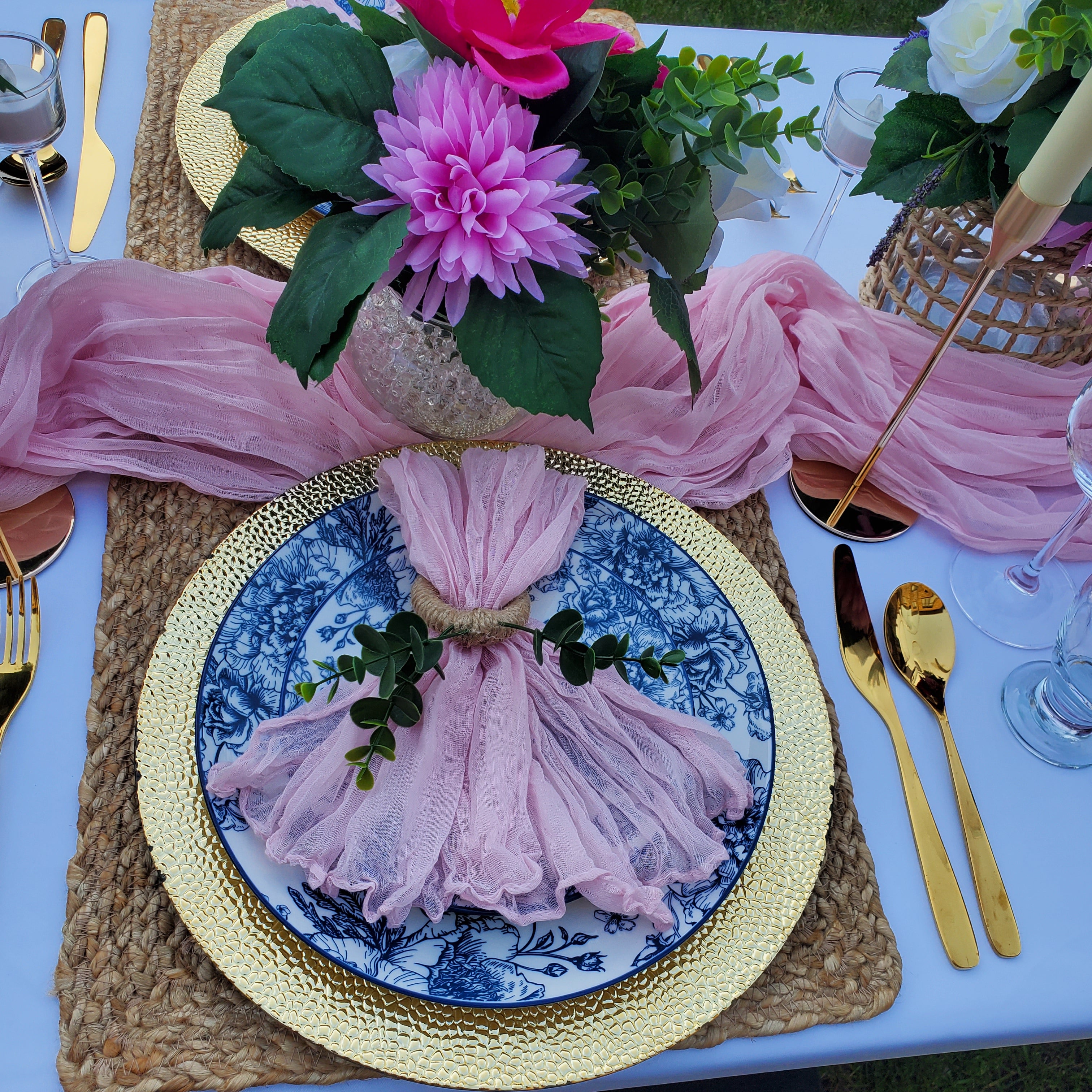 A styled table setting featuring a pink tulle napkin tied with twine and placed on a blue-and-white patterned plate over a gold charger plate, with a matching pink tulle centerpiece and floral arrangement above.