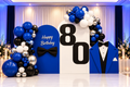 An 80th birthday party backdrop setup featuring a large white arch with black '80' numbers, blue and white balloon garlands, a blue 'Happy Birthday' sign with a black bow tie, and a blue tuxedo cutout on the right.