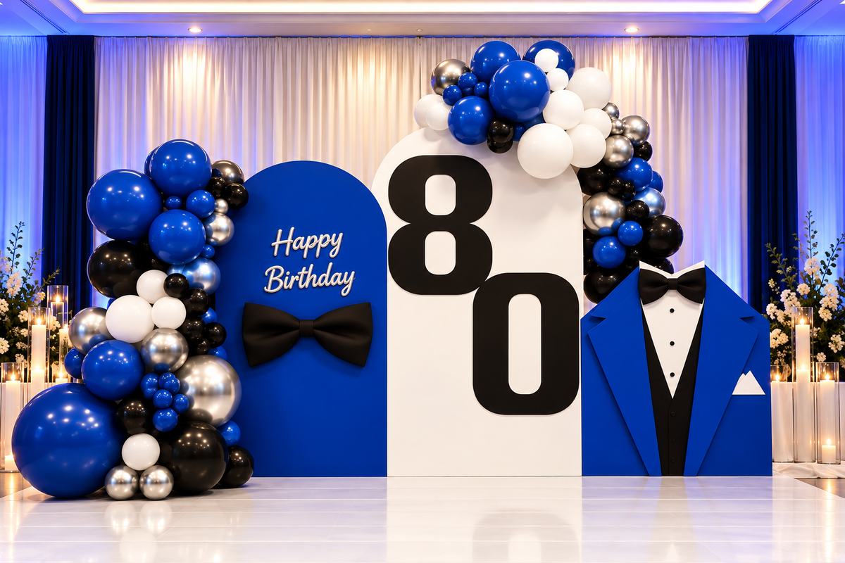 An 80th birthday party backdrop setup featuring a large white arch with black '80' numbers, blue and white balloon garlands, a blue 'Happy Birthday' sign with a black bow tie, and a blue tuxedo cutout on the right.
