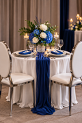 A styled round wedding table with a white tablecloth, two white chairs with chrome frames, a blue floral centerpiece, blue napkins, and a dark blue fabric runner draped over the front of the table.